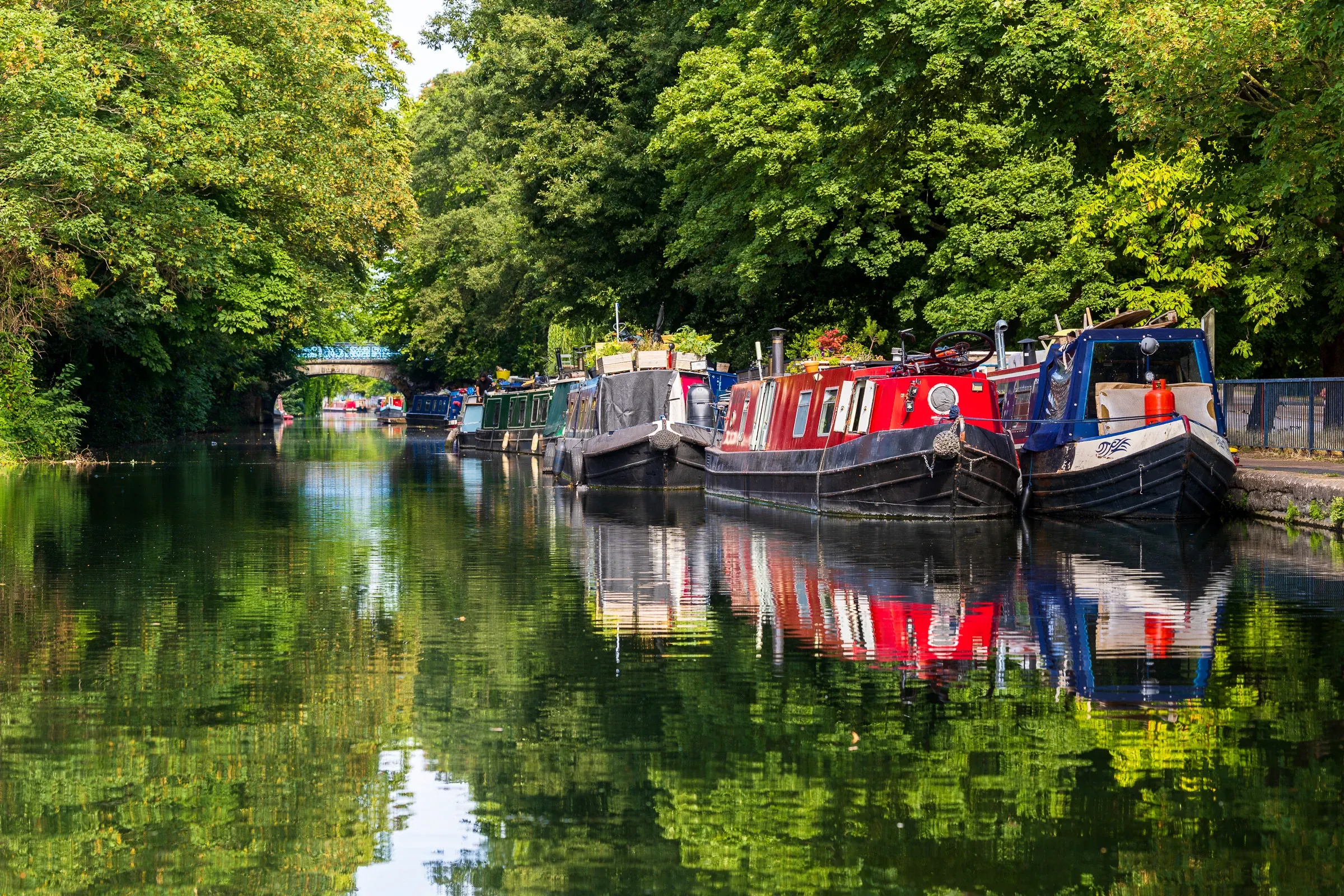 Narrowboats moored along a tree-lined canal, a stone bridge in the distance, reflections in still water