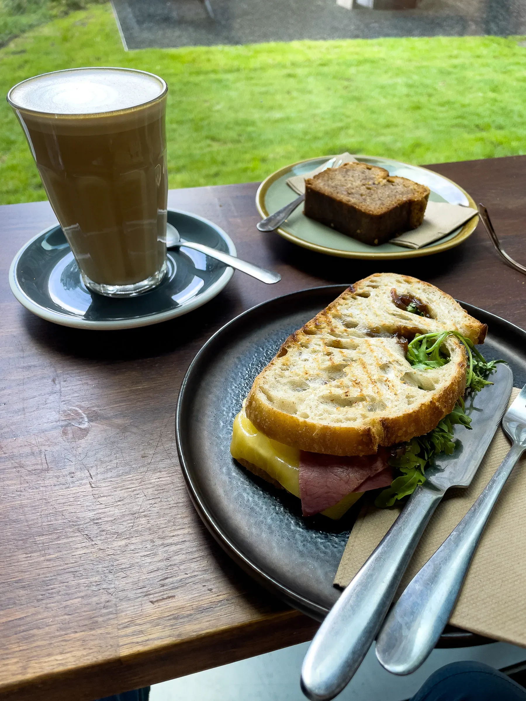 A latte, a slice of banana bread and a ham and cheese toastie on a café table by a window overlooking a lawn