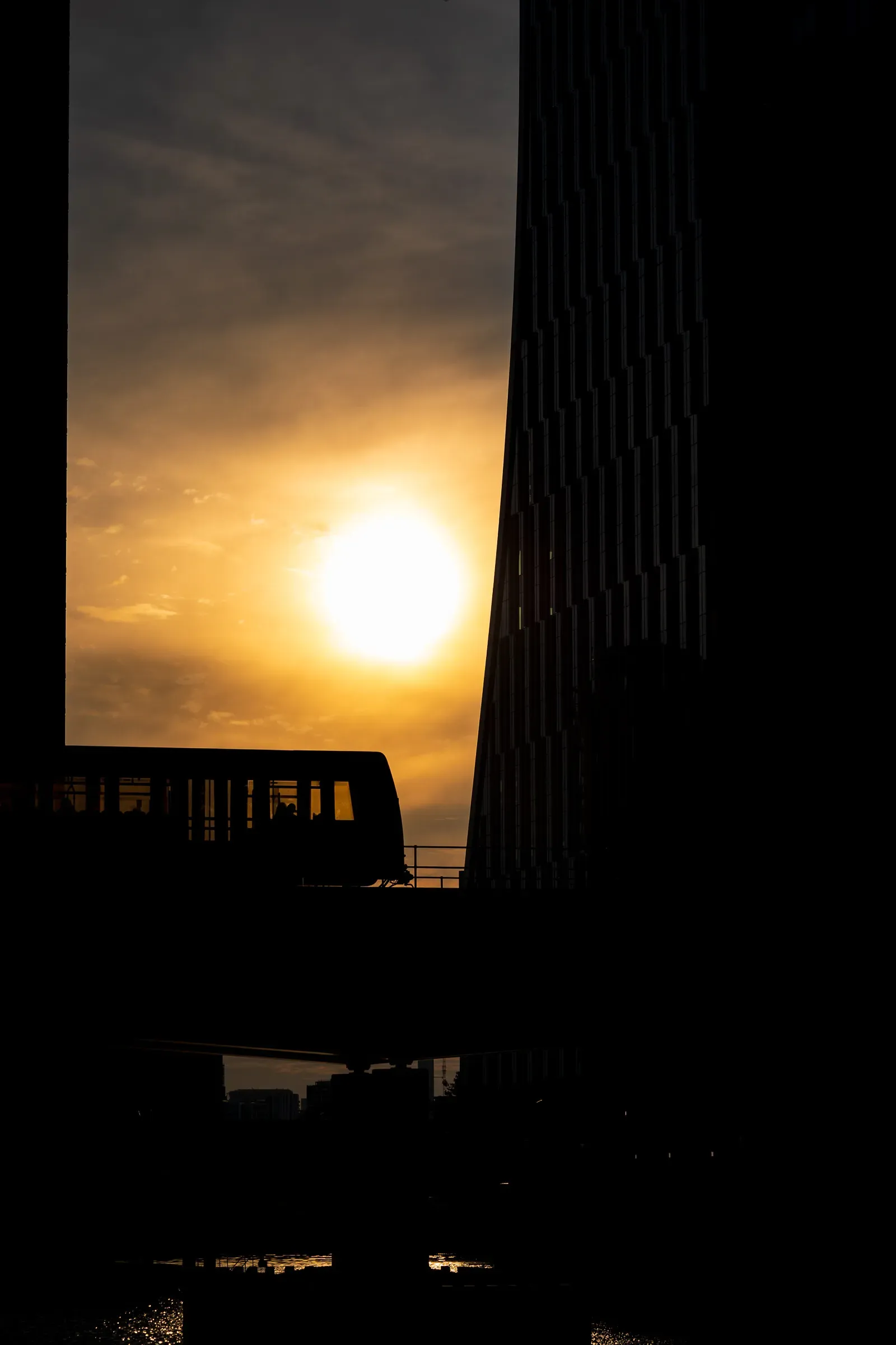A train crossing a bridge in silhouette against a setting sun, a dark angular tower beside it