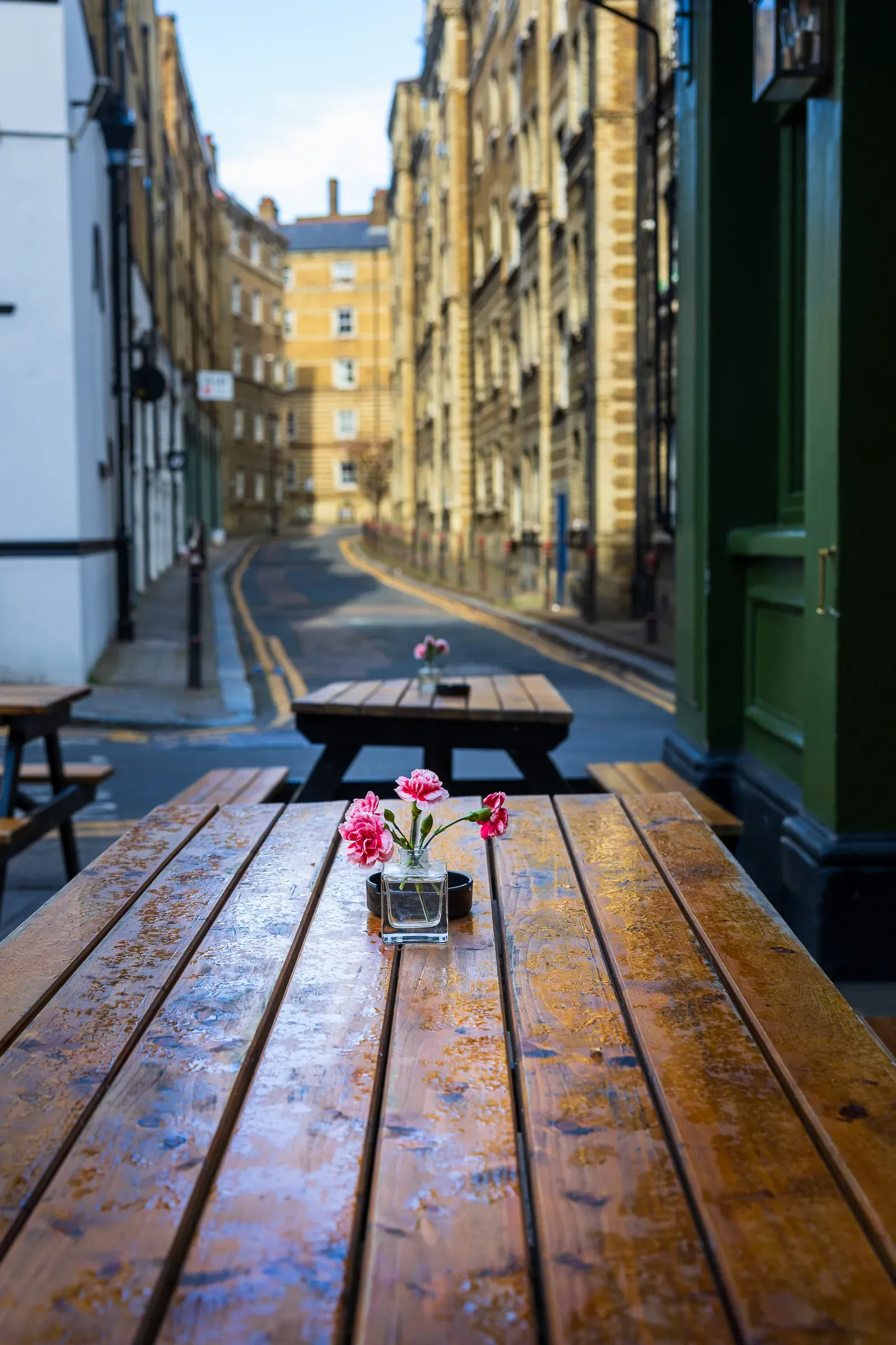 Empty pub garden picnic tables with small vases of pink carnations, a cobbled lane and warehouse buildings beyond