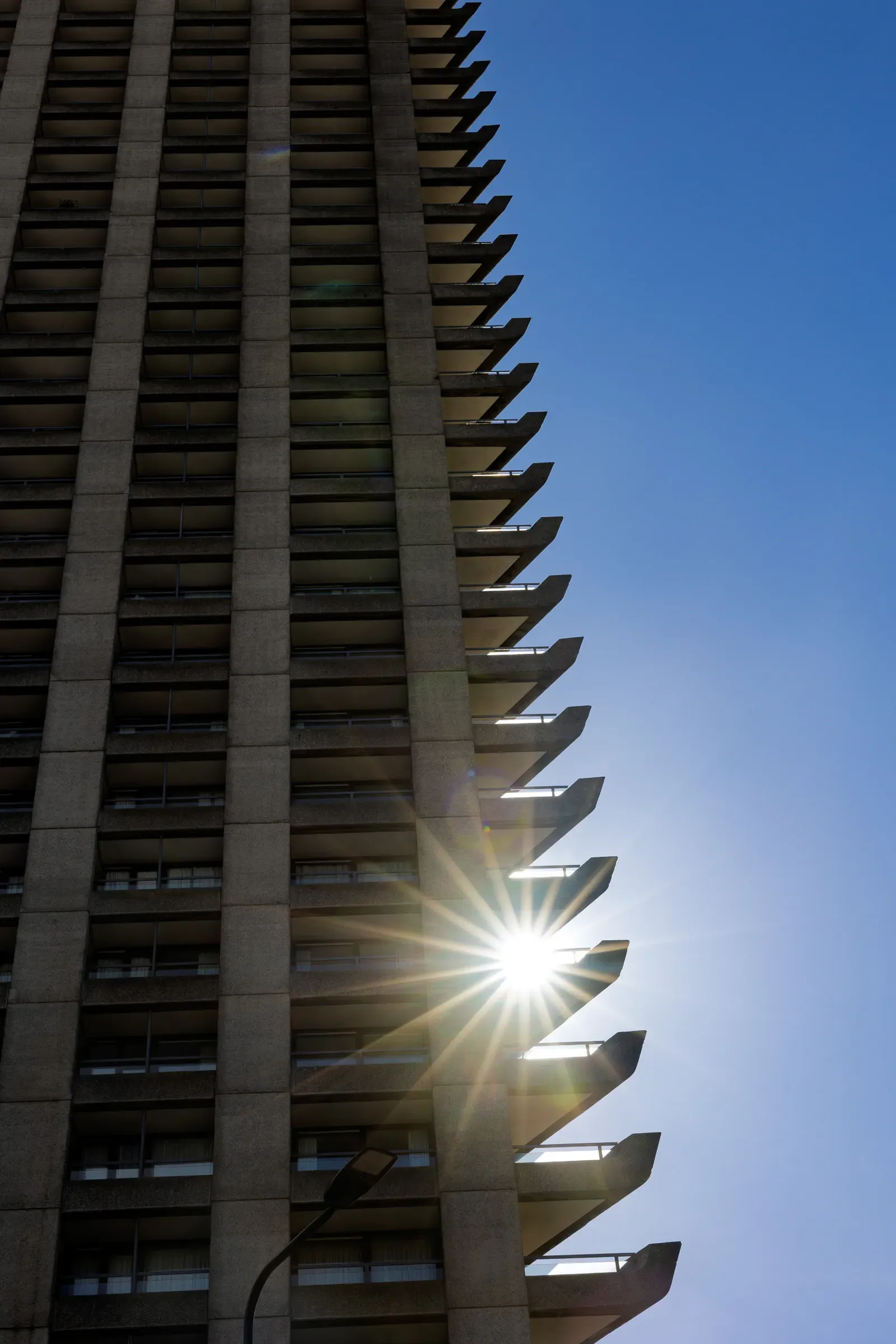 Sun flaring between the angular concrete balconies of a Barbican tower against a clear blue sky