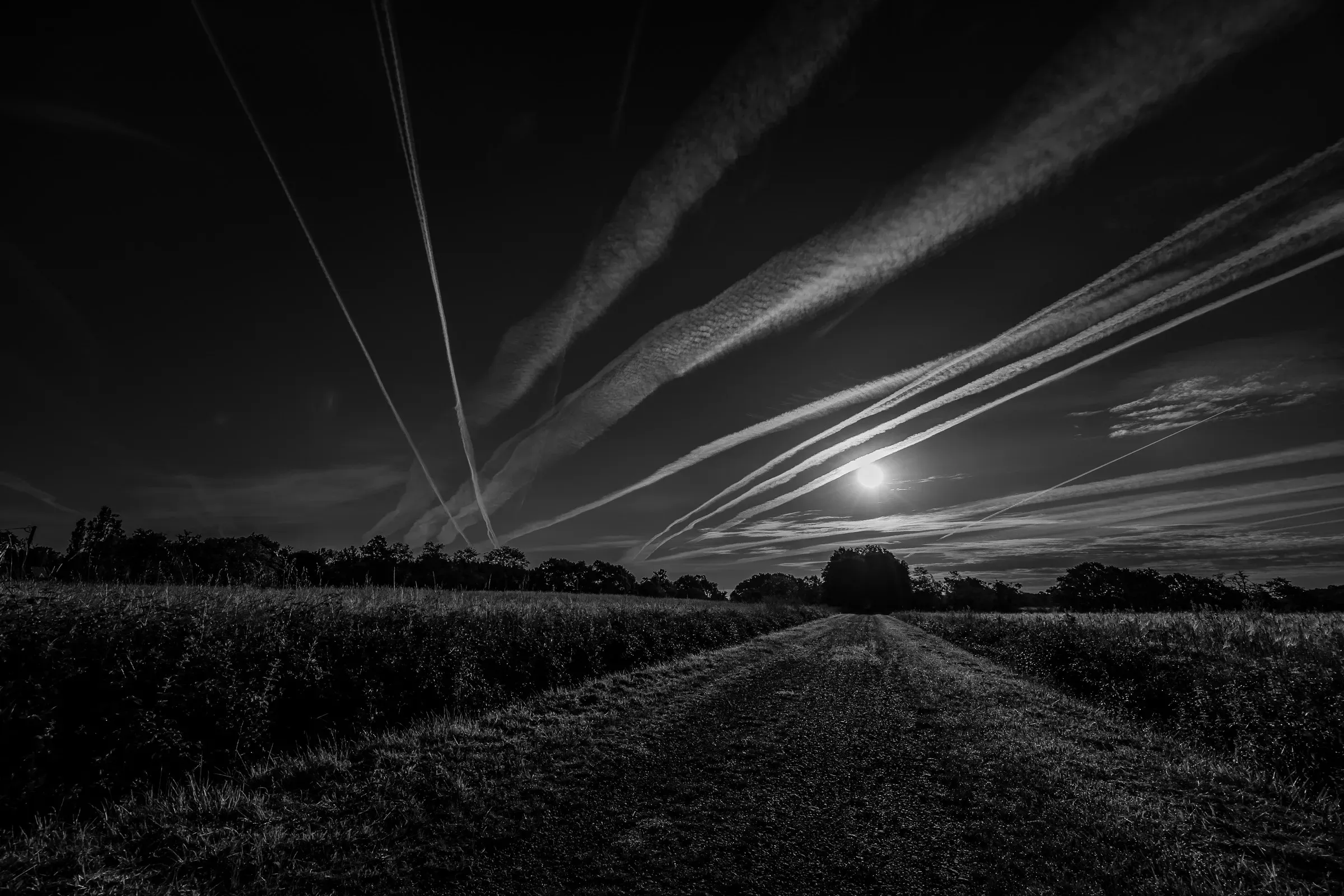 Gravel farm track bisecting flat fields at dusk, a pale moon sitting low above the treeline, sky filled with contrails radiating outward from the horizon