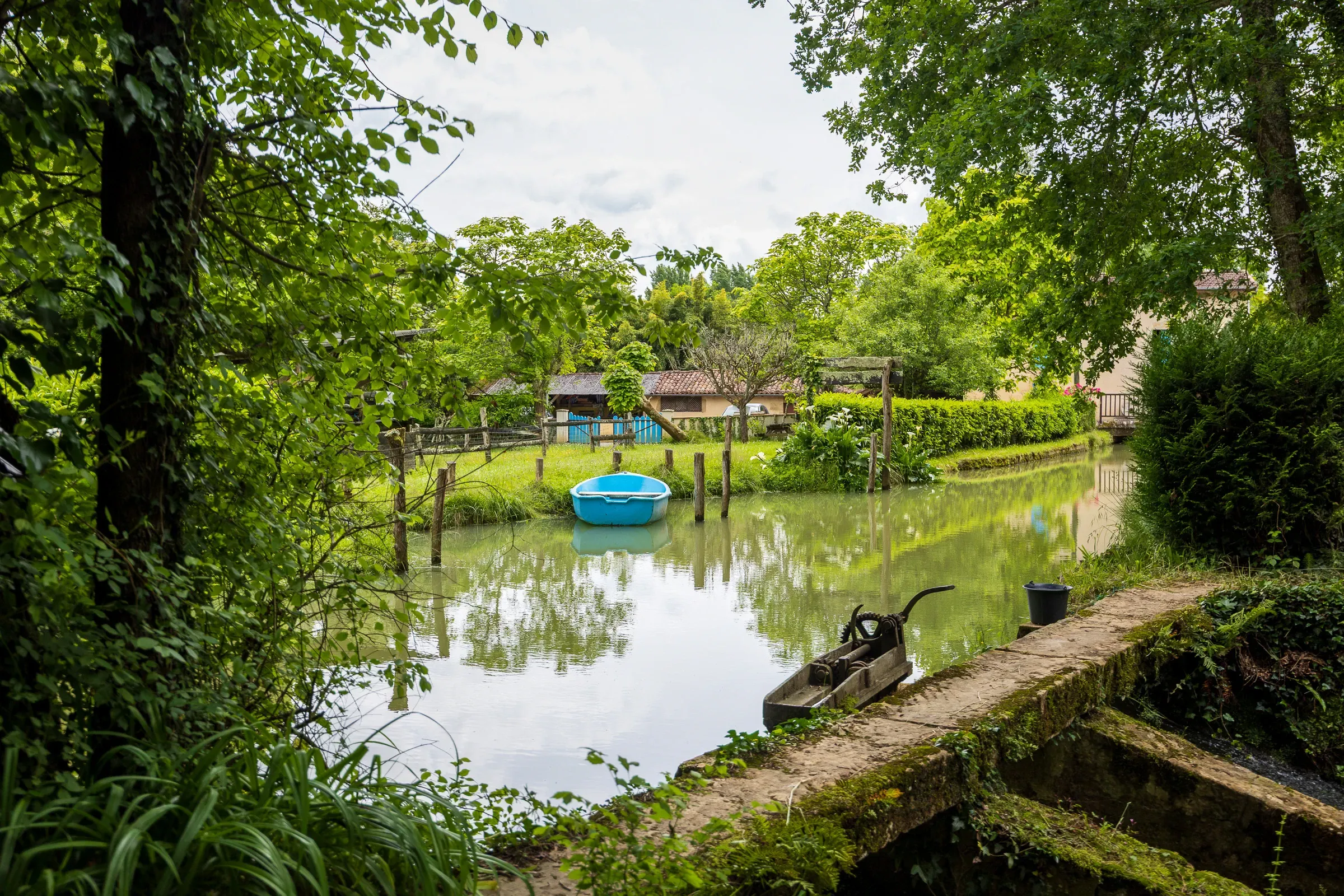Blue plastic rowboat moored in a green canal pond, framed by trees, with a farmhouse beyond