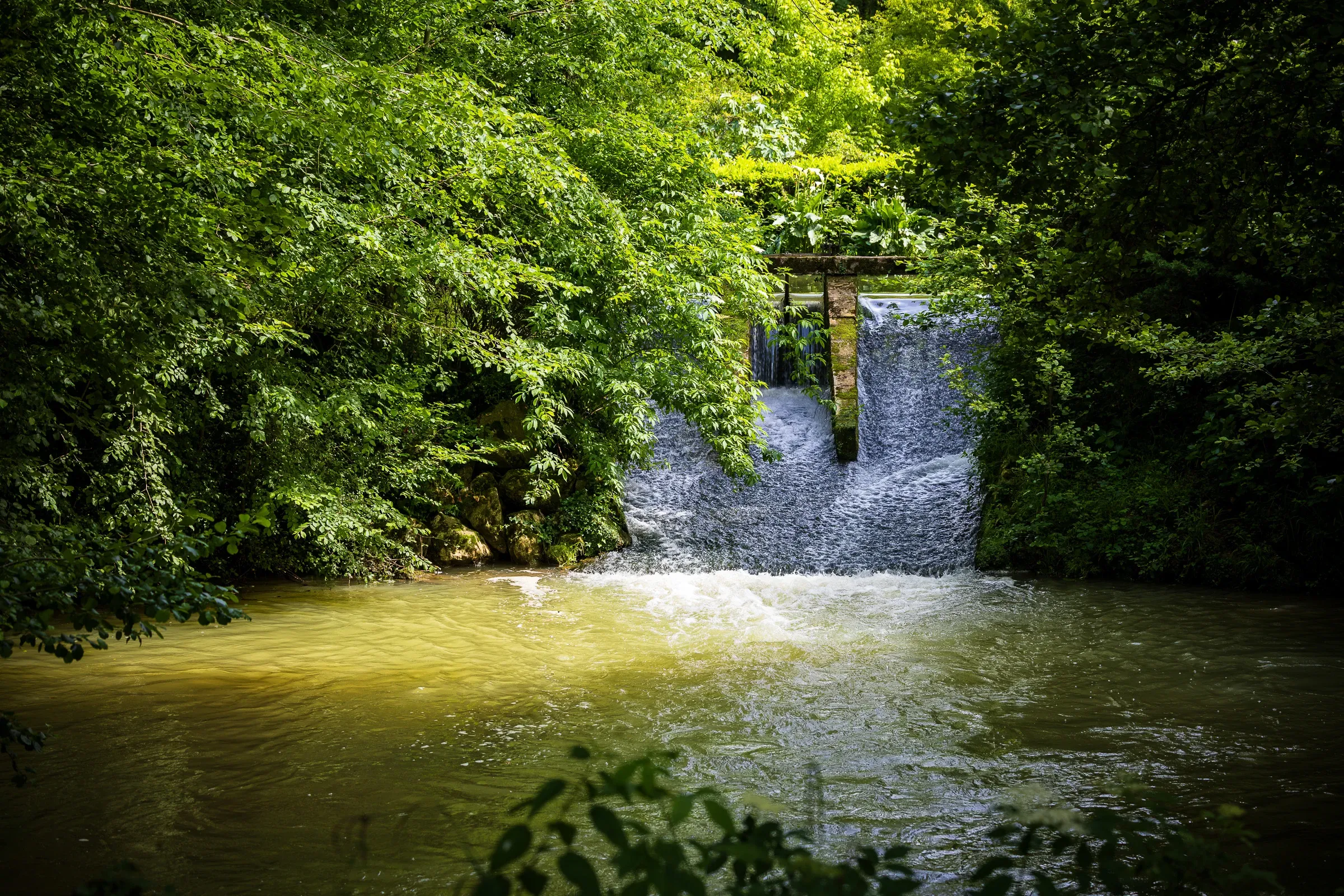 Water rushing through a stone sluice gate flanked by dense green trees