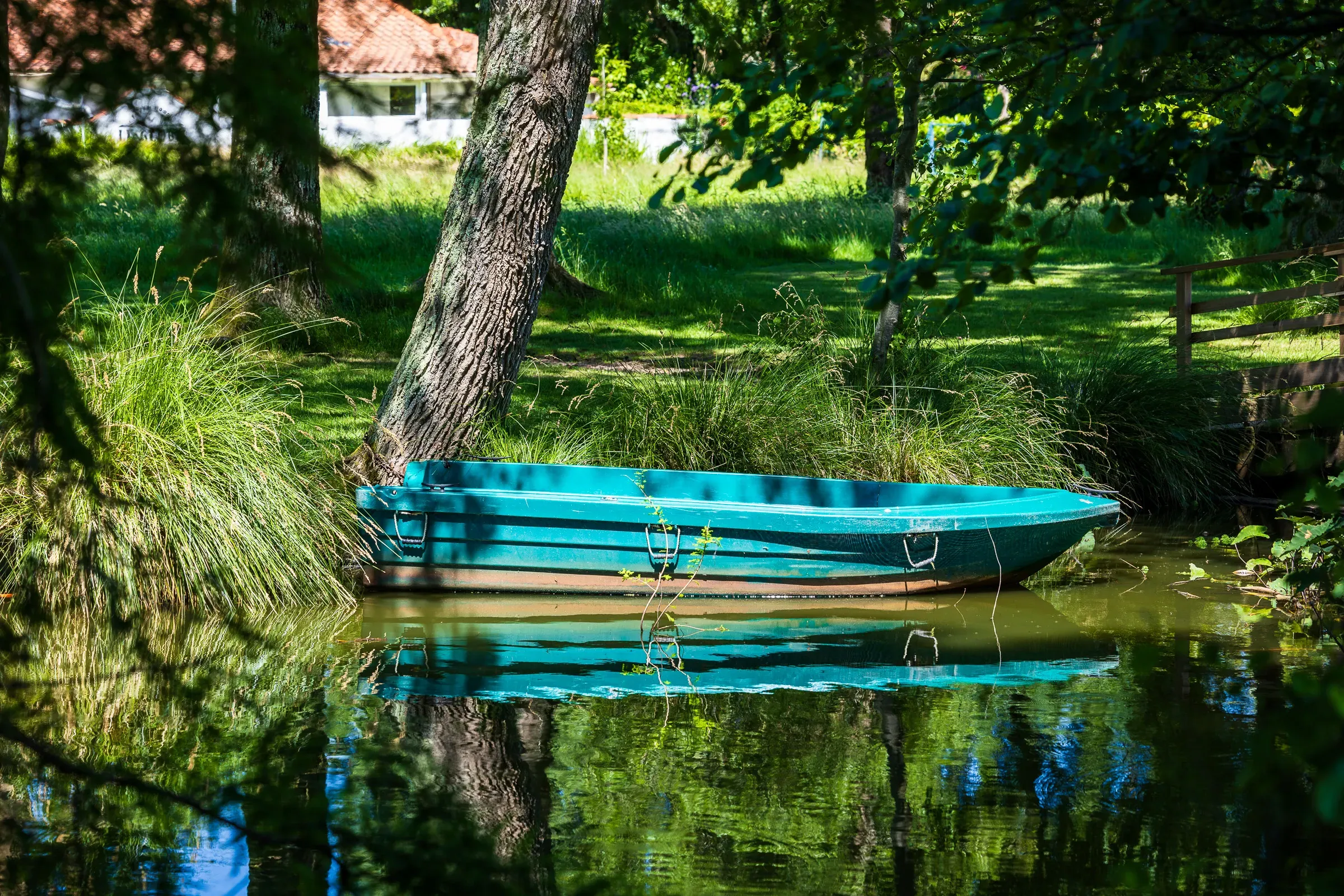 Turquoise rowboat moored in a shaded green canal, reflected sharply in calm water