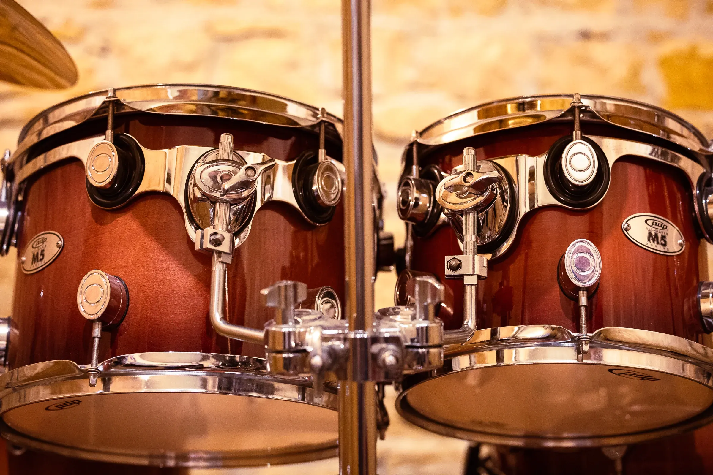Two PDP M5 tom-toms in close-up, dark red sunburst finish, chrome lugs and tension rods, warm stone wall behind, Lyon