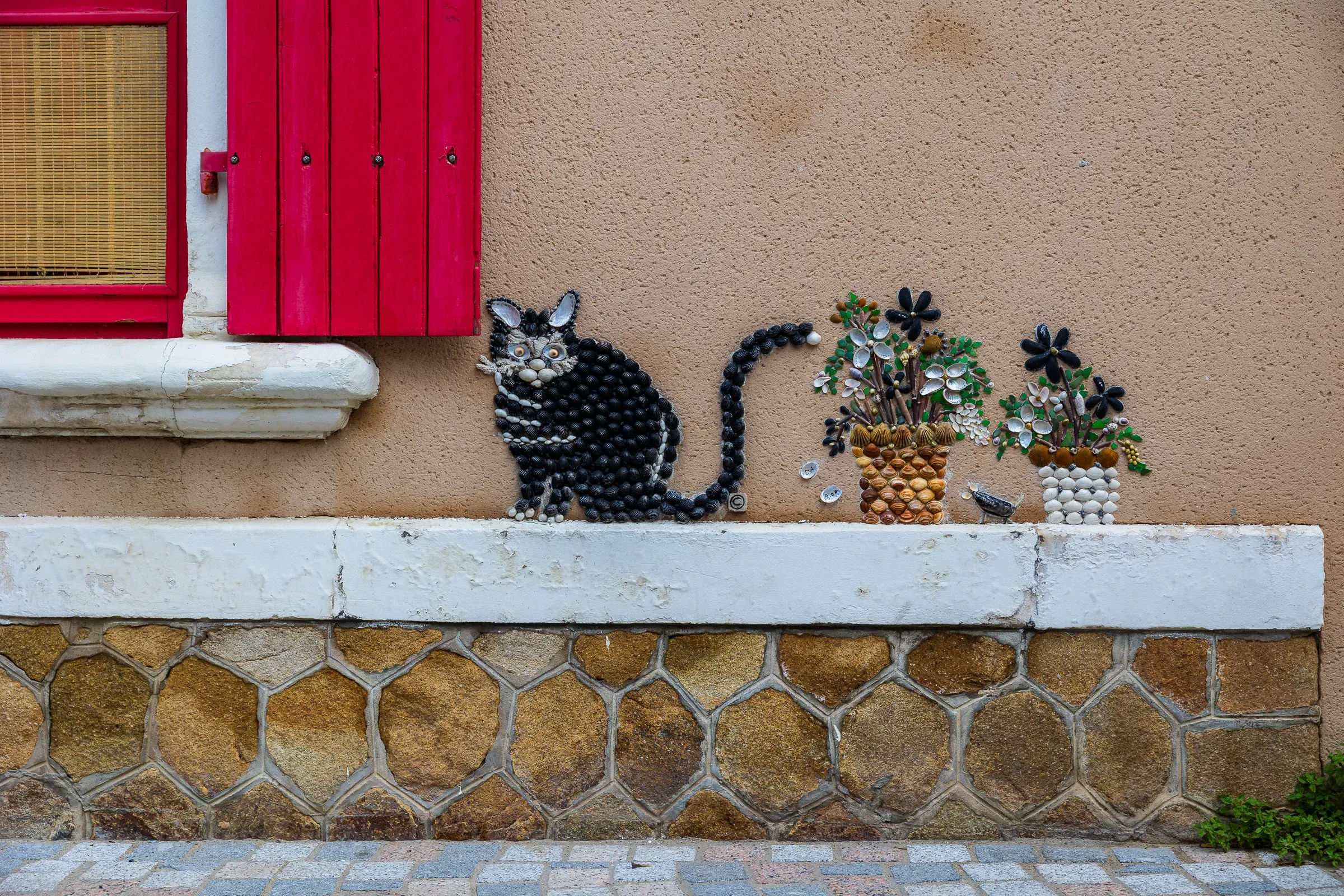 Black cat and flower pot mosaic made of shells on a rendered wall with red shutters