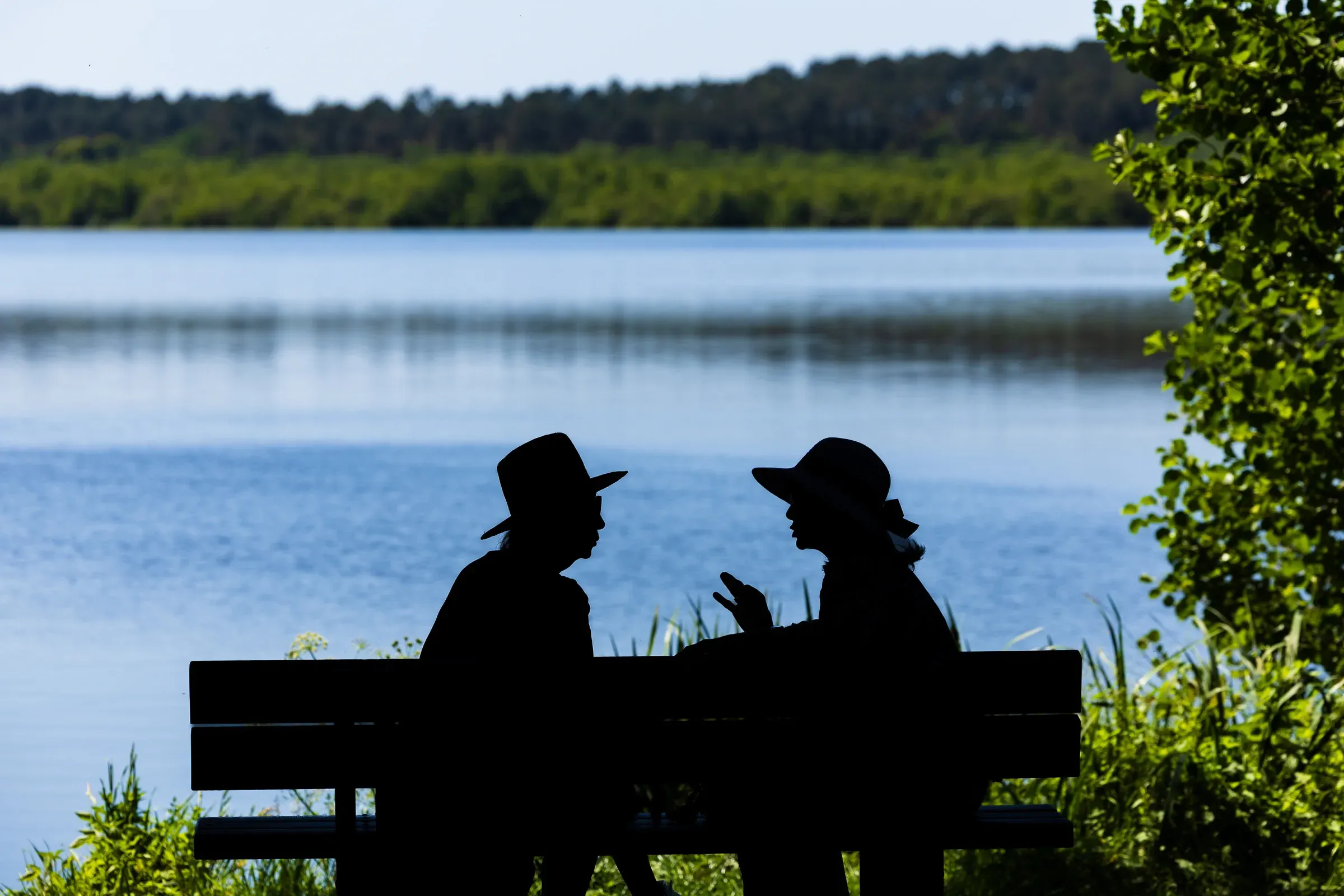 Two silhouetted figures in hats sitting on a bench by a lake, deep in conversation