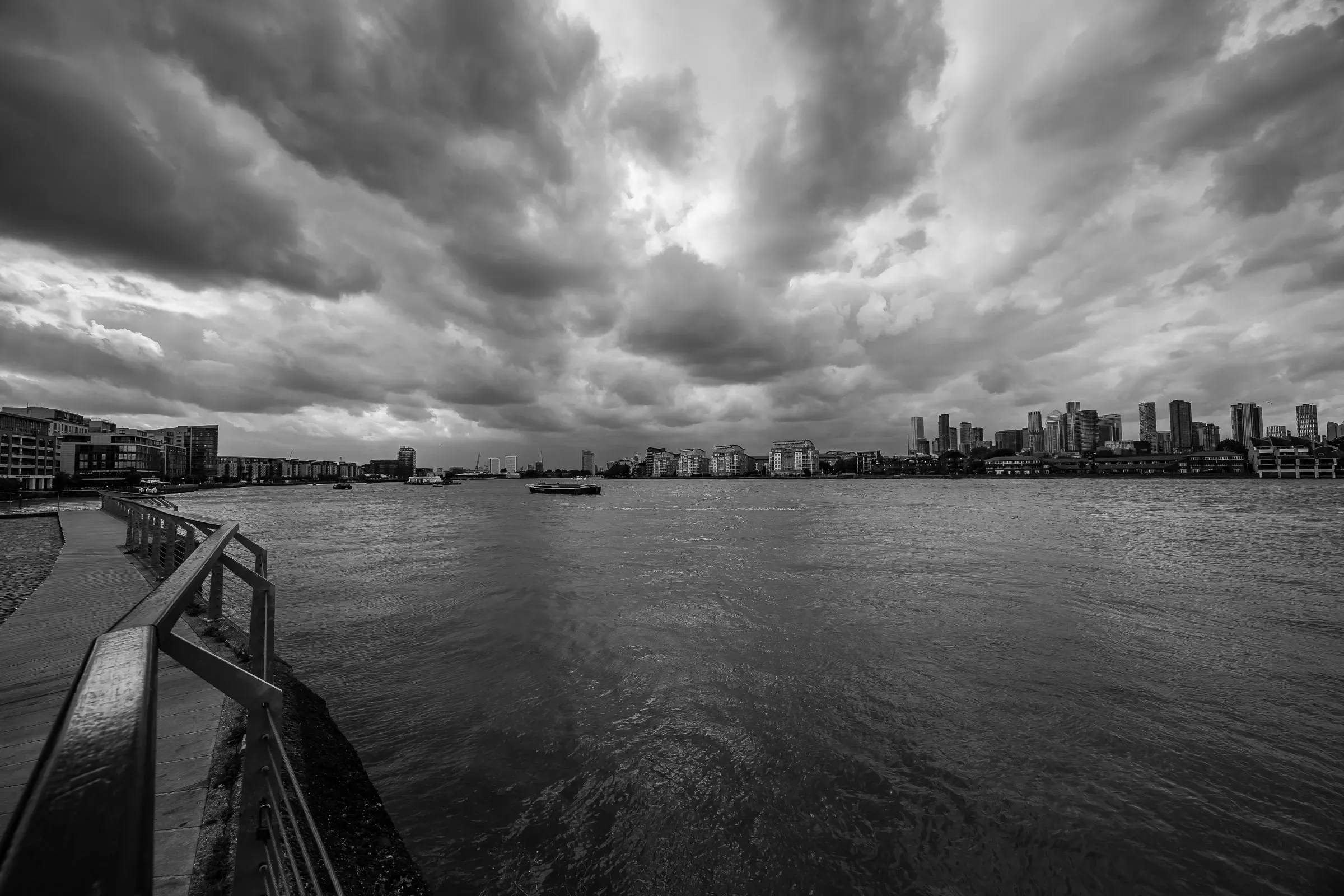 Storm clouds over the Thames, a wooden pier and railing in the foreground, the Canary Wharf skyline across the water, black and white